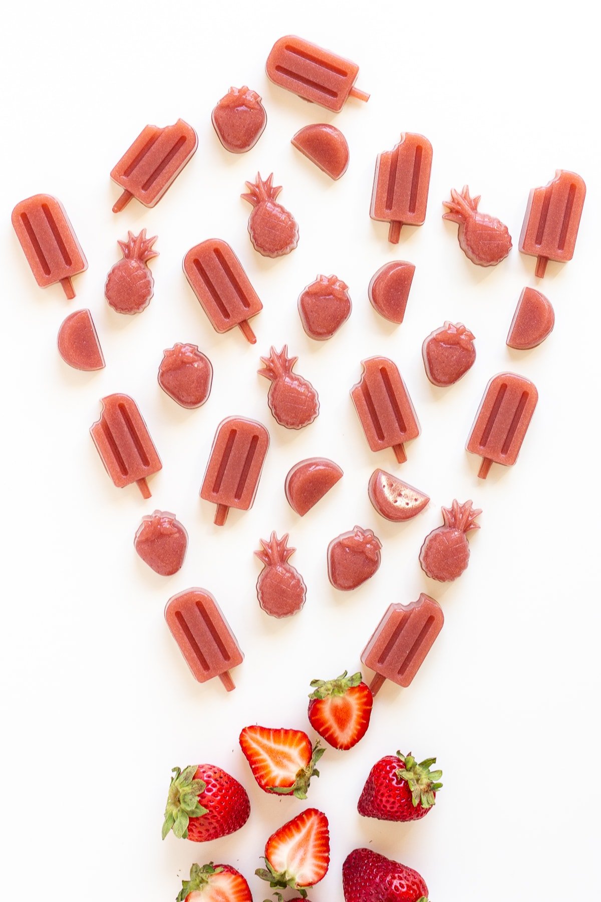 Variety of homemade strawberry gummies on a white background in the shape of popsicles, pineapples, watermelon slices and strawberries.