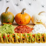 A Halloween themed taco board featuring three carved pumpkins "puking" guacamole, salsa and sour cream.