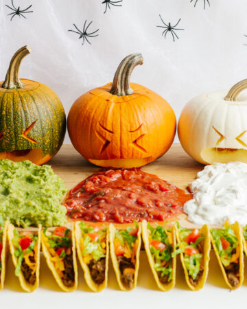 A Halloween themed taco board featuring three carved pumpkins "puking" guacamole, salsa and sour cream.