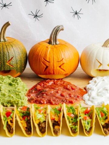 A Halloween themed taco board featuring three carved pumpkins "puking" guacamole, salsa and sour cream.