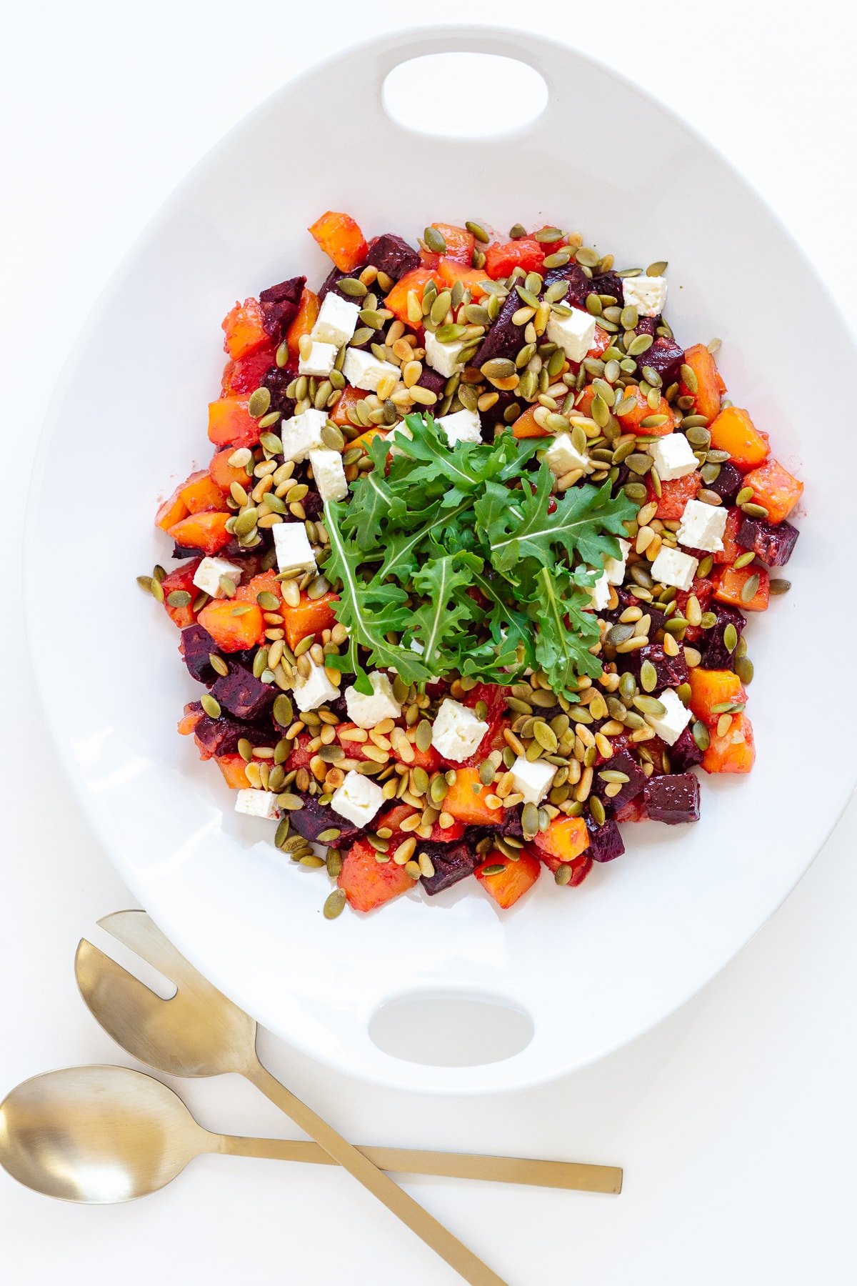 Pumpkin and beetroot salad in a white serving dish with matte gold coloured salad servers.