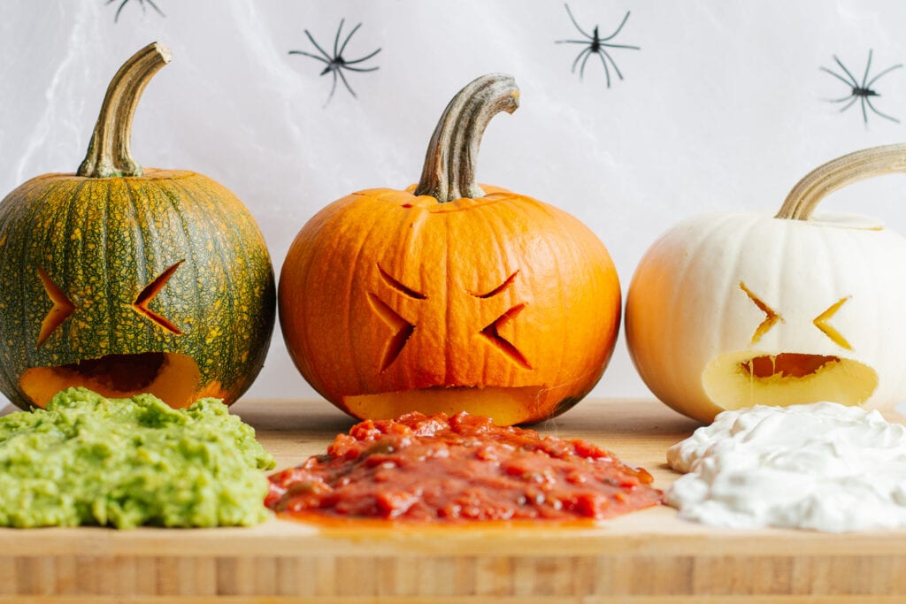 Three small carved pumpkins on a wooden board that look like they are throwing up guacamole, salsa and sour cream.