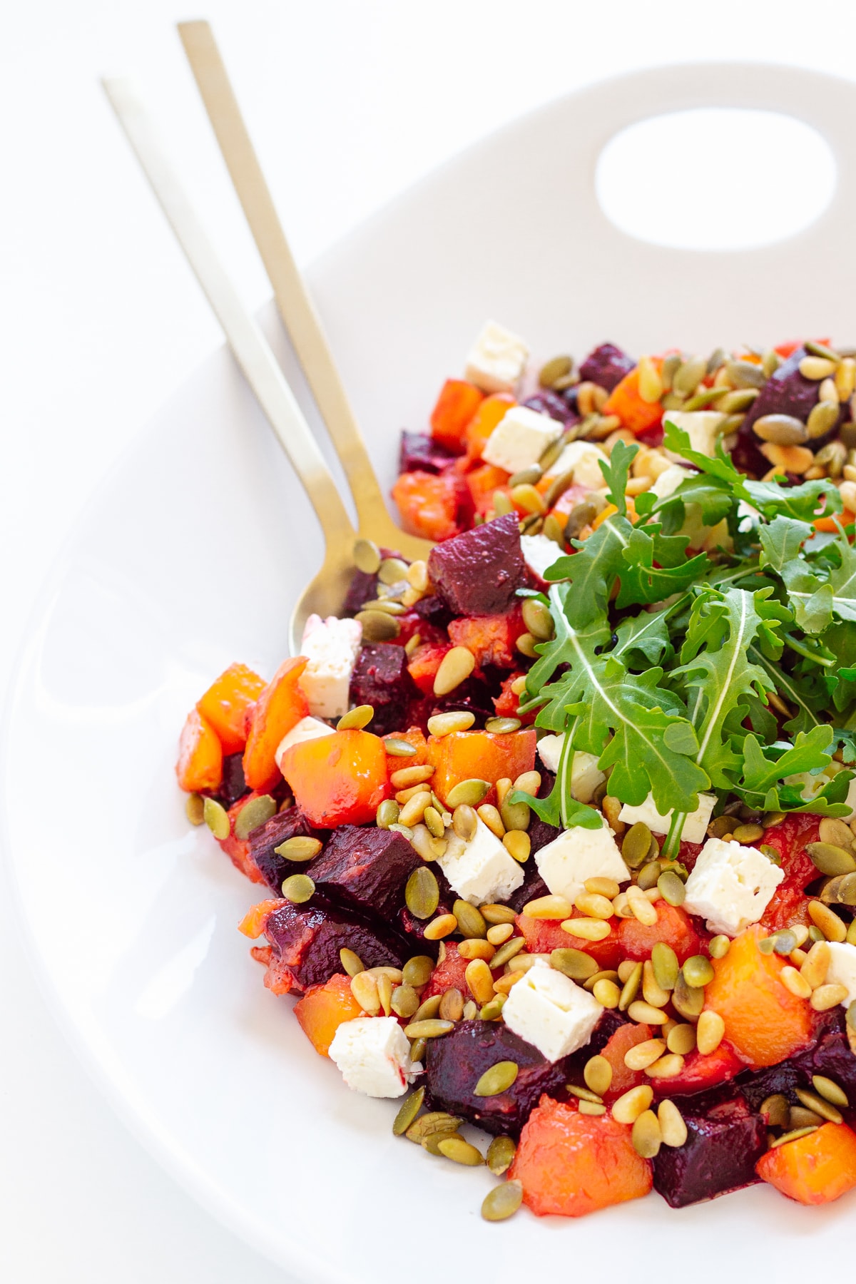 A fall pumpkin and beet salad in a white serving bowl with gold salad servers.