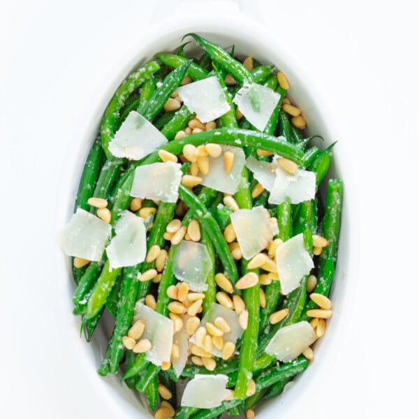 Garlic butter green beans in a white oval serving dish, garnished with pine nuts and parmesan shavings.