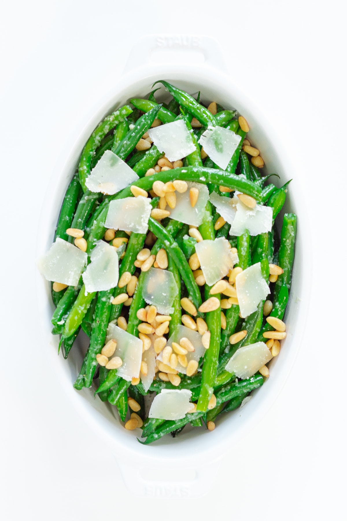 Sauteed garlic butter green beans in a white serving dish with parmesan shavings and pine nuts on top.