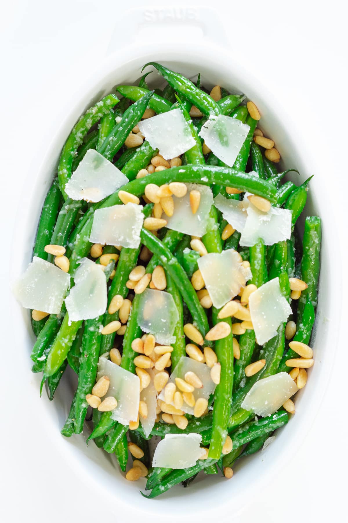 White serving dish containing garlic green beans garnished with parmesan and pine nuts.