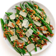 Garlic butter parmesan green beans on a white serving plate garnished with pine nuts, parmesan shavings and crispy fried shallots.