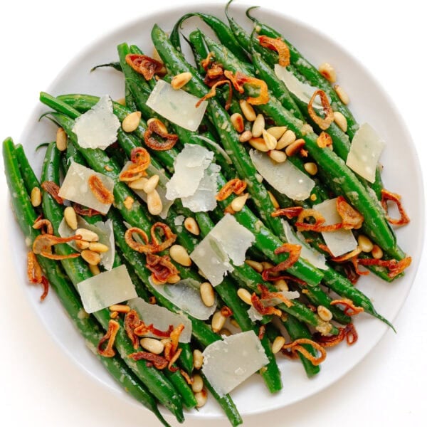 Garlic butter parmesan green beans on a white serving plate garnished with pine nuts, parmesan shavings and crispy fried shallots.