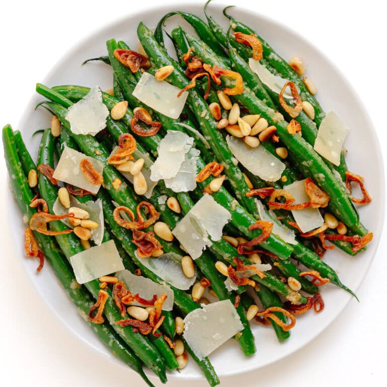 Garlic butter parmesan green beans on a white serving plate garnished with pine nuts, parmesan shavings and crispy fried shallots.