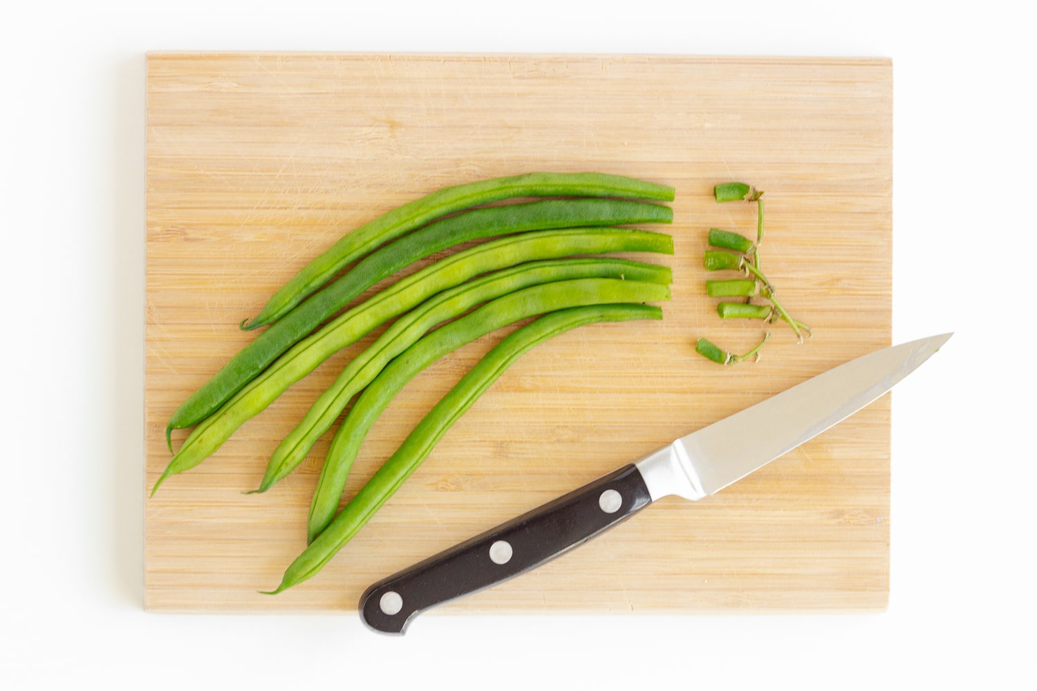 Green beans with ends trimmed off and a knife sitting on a wood cutting board.