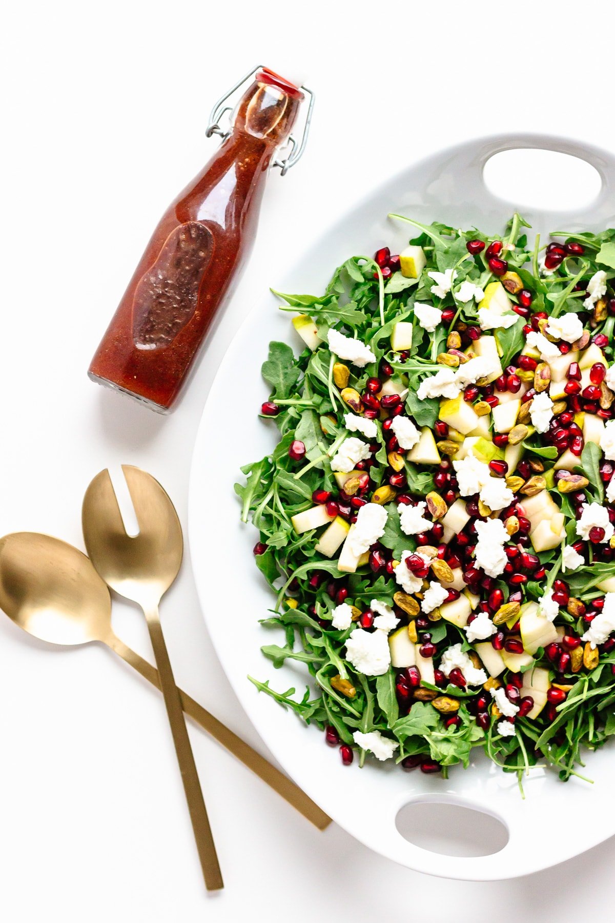 Overhead photo of a platter of winter holiday salad, gold coloured salad servers and a bottle of pomegranate vinaigrette.