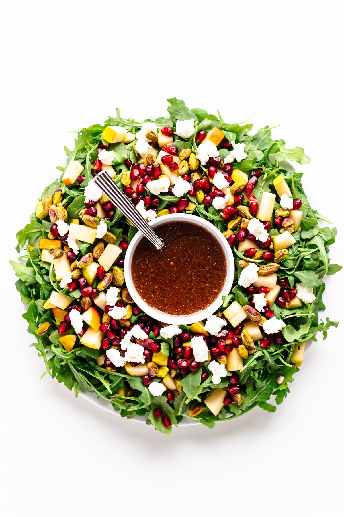 Holiday salad arranged in the shape of a Christmas wreath with a bowl of pomegranate vinaigrette in the center.