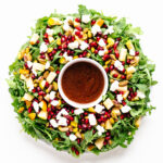 Winter Holiday Salad (arugula, pear, pomegranate arils, pistachios, goat cheese) arranged in a Christmas wreath shape with small bowl of pomegranate vinaigrette in the center.