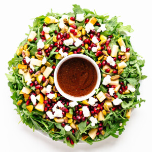 Winter Holiday Salad (arugula, pear, pomegranate arils, pistachios, goat cheese) arranged in a Christmas wreath shape with small bowl of pomegranate vinaigrette in the center.