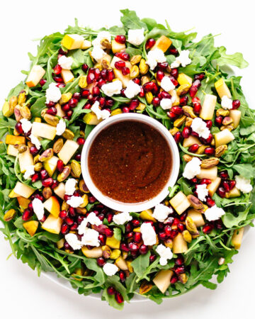 Winter Holiday Salad (arugula, pear, pomegranate arils, pistachios, goat cheese) arranged in a Christmas wreath shape with small bowl of pomegranate vinaigrette in the center.