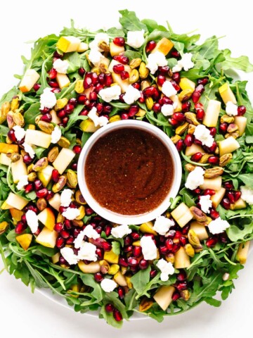 Winter Holiday Salad (arugula, pear, pomegranate arils, pistachios, goat cheese) arranged in a Christmas wreath shape with small bowl of pomegranate vinaigrette in the center.