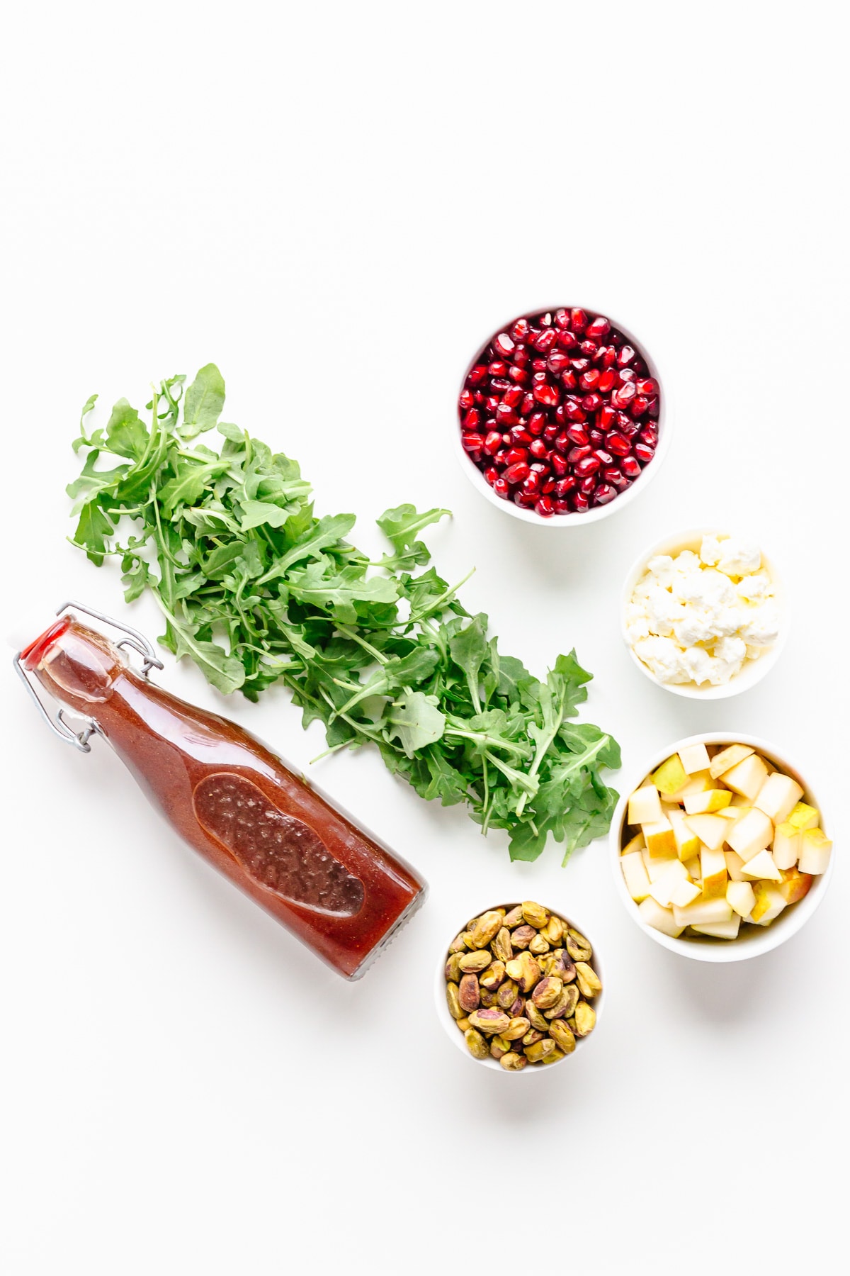 Overhead photo of ingredients needed for winter holiday salad recipe: arugula, pomegranate arils, goat cheese, pear, pistachios and pomegranate vinaigrette.