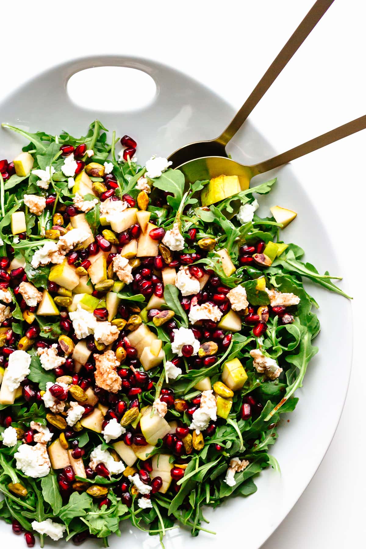 White platter of winter holiday salad (baby arugula, pear, pistachios, pomegranate, goat cheese) with gold salad servers.
