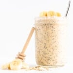 Jar of banana overnight oats with honey dipper leaning against it and banana slices and rolled oats scattered next to it.