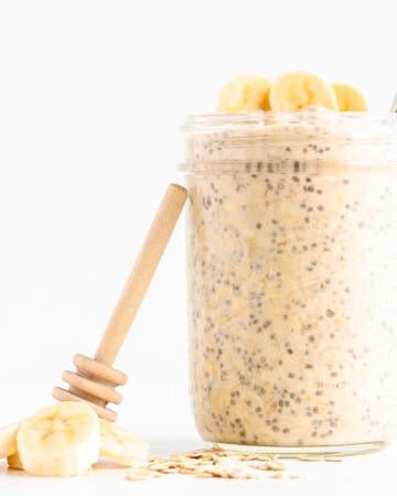 Jar of banana overnight oats with honey dipper leaning against it and banana slices and rolled oats scattered next to it.