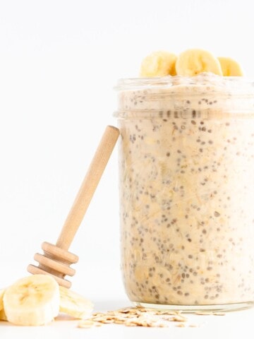 Jar of banana overnight oats with honey dipper leaning against it and banana slices and rolled oats scattered next to it.