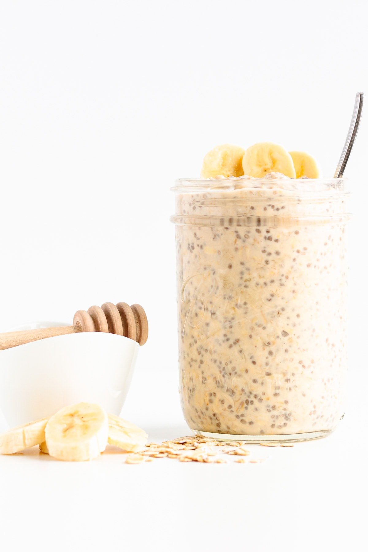 Mason jar of banana overnight oats with honey, banana slices and some rolled oats scattered next to it.