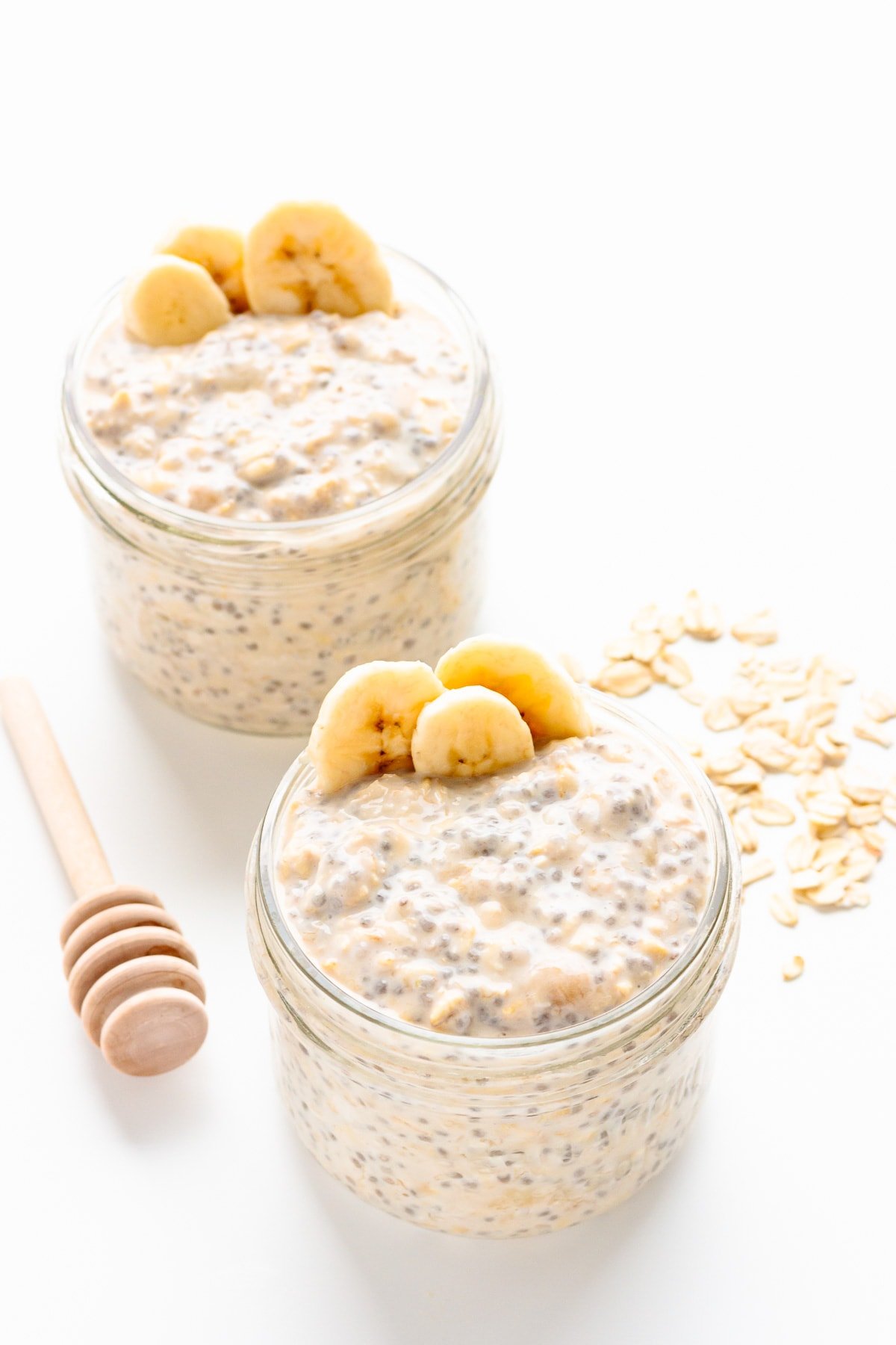Two jars of banana overnight oats garnished with banana slices with a honey dipper and some rolled oats beside them.