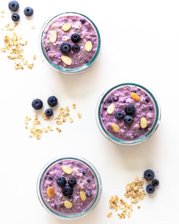 Three containers of blueberry overnight oats with blueberries and rolled oats scattered beside them.