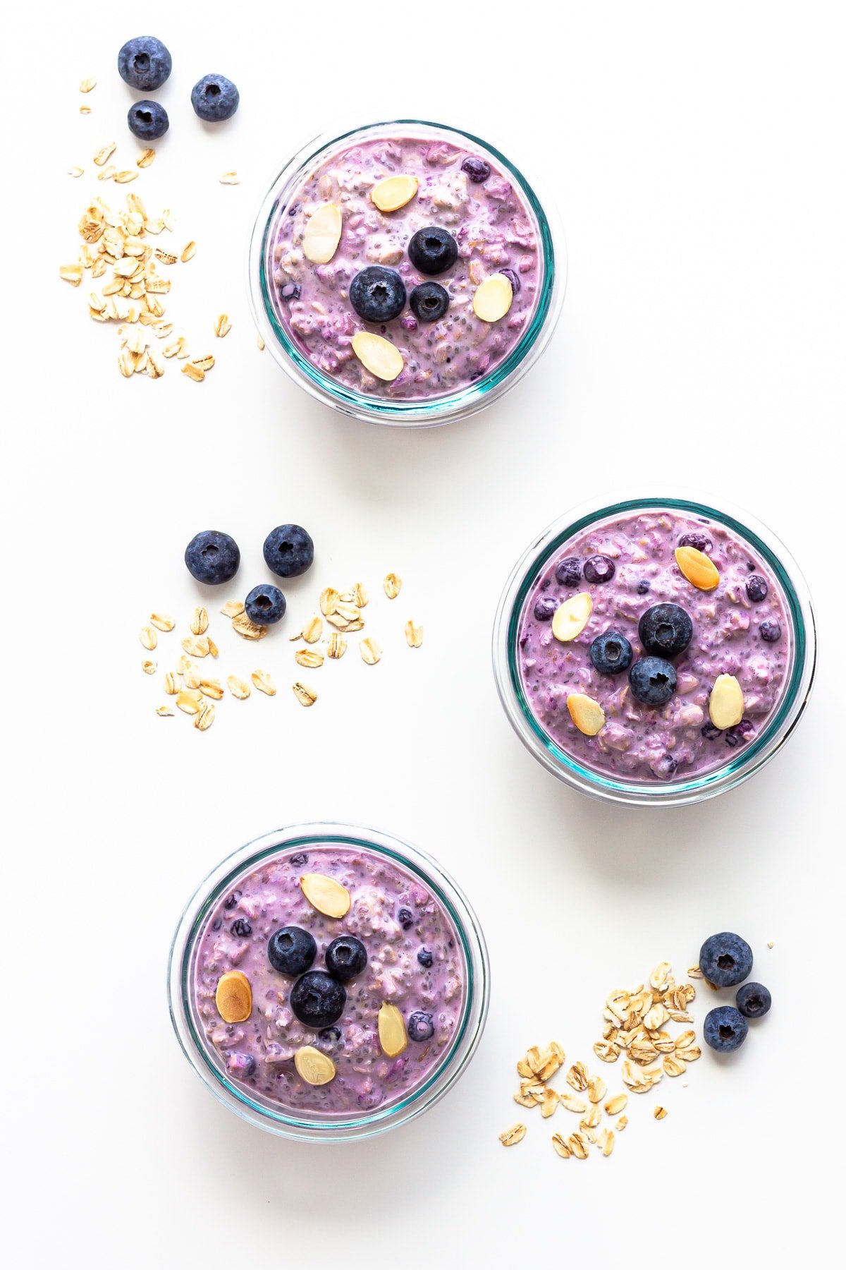 Overhead photo of three containers of blueberry overnight oats with blueberries and rolled oats scattered beside them.