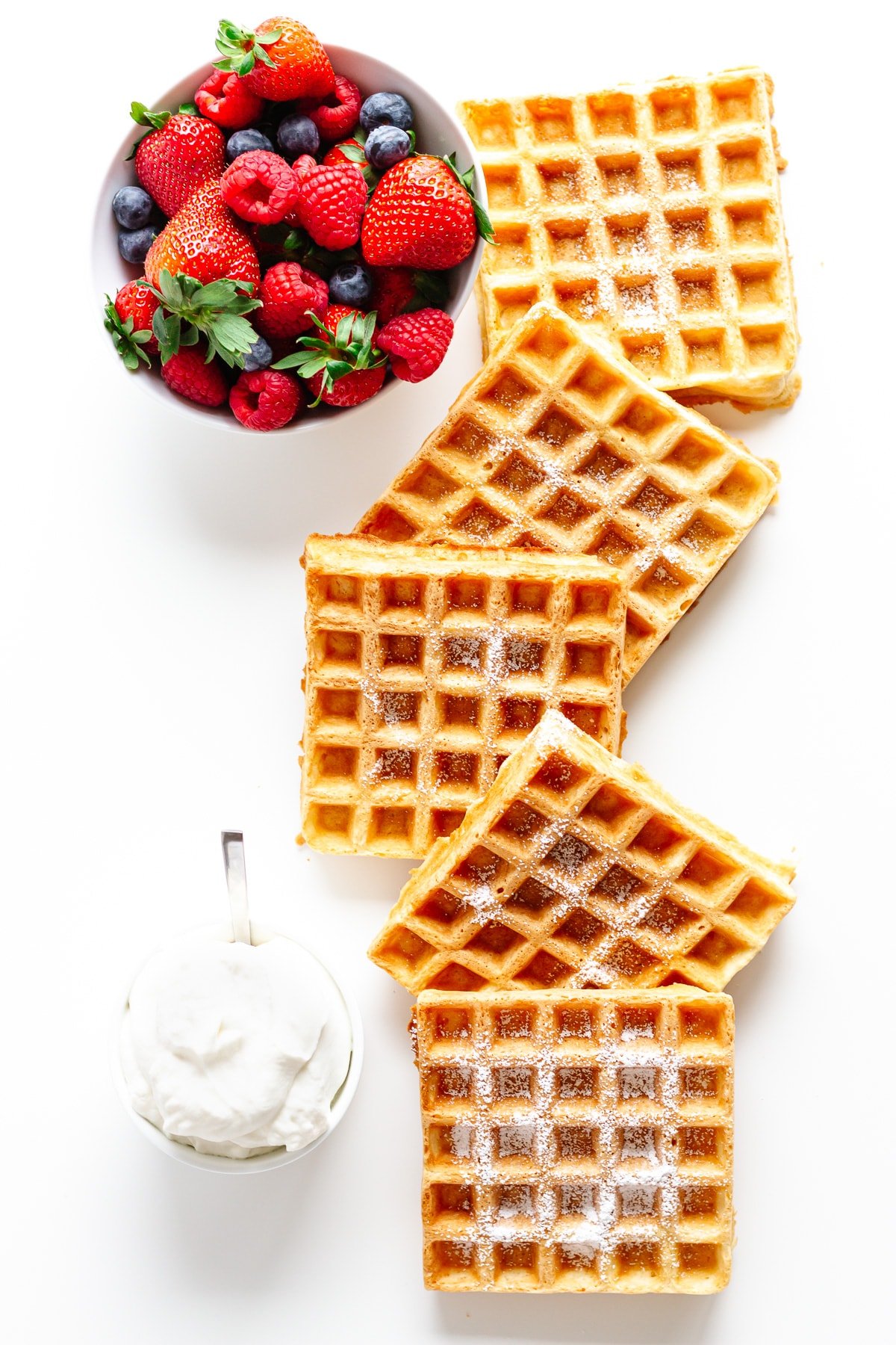 Overhead photo of square shaped waffles, bowl of berries and whipped cream on a white background.