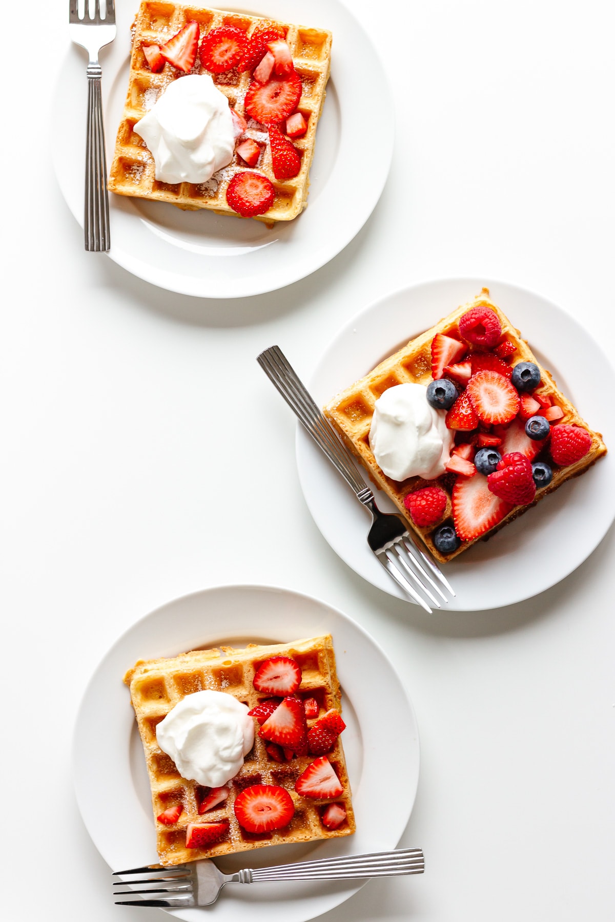 Three waffles on white plates with a fork and topped with berries and whipped cream.