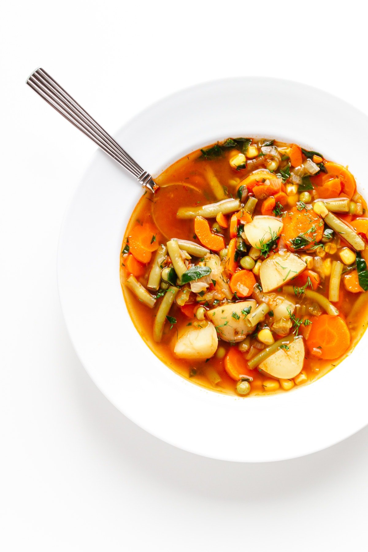 Vegetable soup in white bowl with spoon handle sticking out.