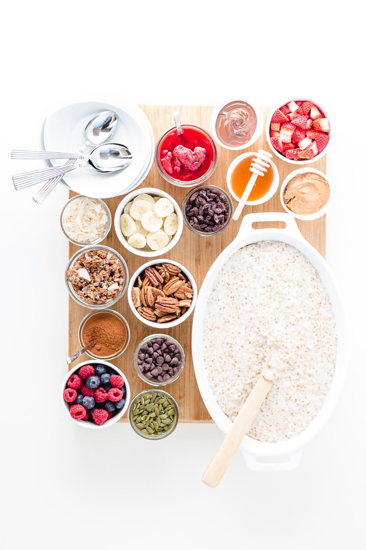 Wooden board topped with a large dish of steel cut oats, variety of small bowls of different toppings and a stack of serving bowls and spoons.