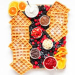 Overhead photo of a waffle breakfast board featuring waffles, bowls of toppings and fresh fruit.