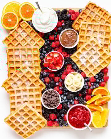 Overhead photo of a waffle breakfast board featuring waffles, bowls of toppings and fresh fruit.