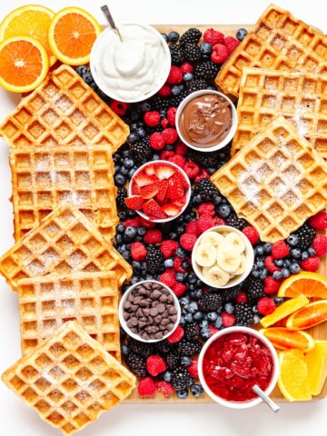 Overhead photo of a waffle breakfast board featuring waffles, bowls of toppings and fresh fruit.