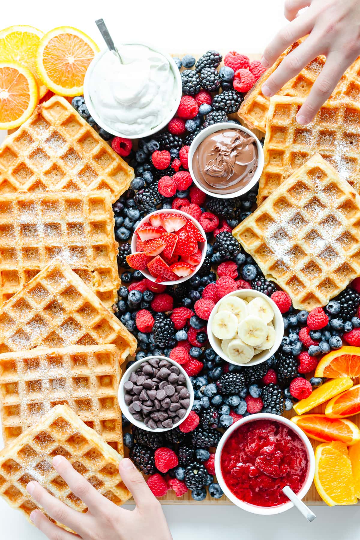 Overhead photo of a waffle breakfast charcuterie board with two children's hands reaching in to grab waffles.