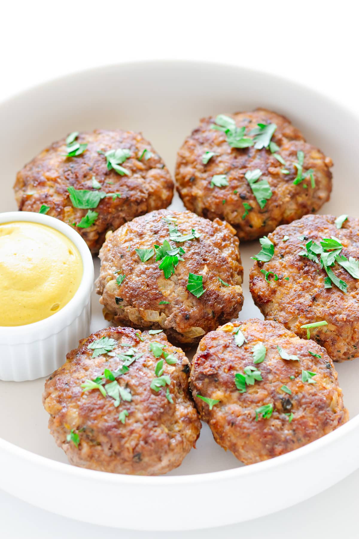 Closeup of six frikadellen in a round white dish with small dish of mustard and garnished with parsley.