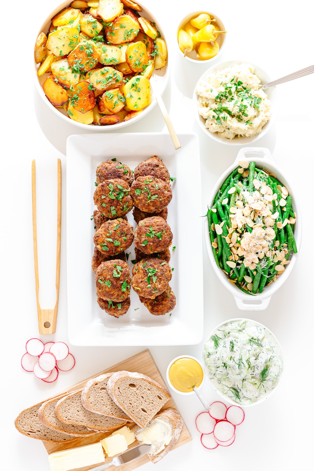 Overhead shot of a dinner table featuring a platter of German frikadellen and a variety of side dishes including pan-fried potatoes, potato salad, green beans, cucumber salad, rye bread, etc.