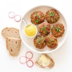 Overhead view of six German frikadellen and small bowl of mustard on a round white serving platter surrounded by some slices of rye bread and radishes.