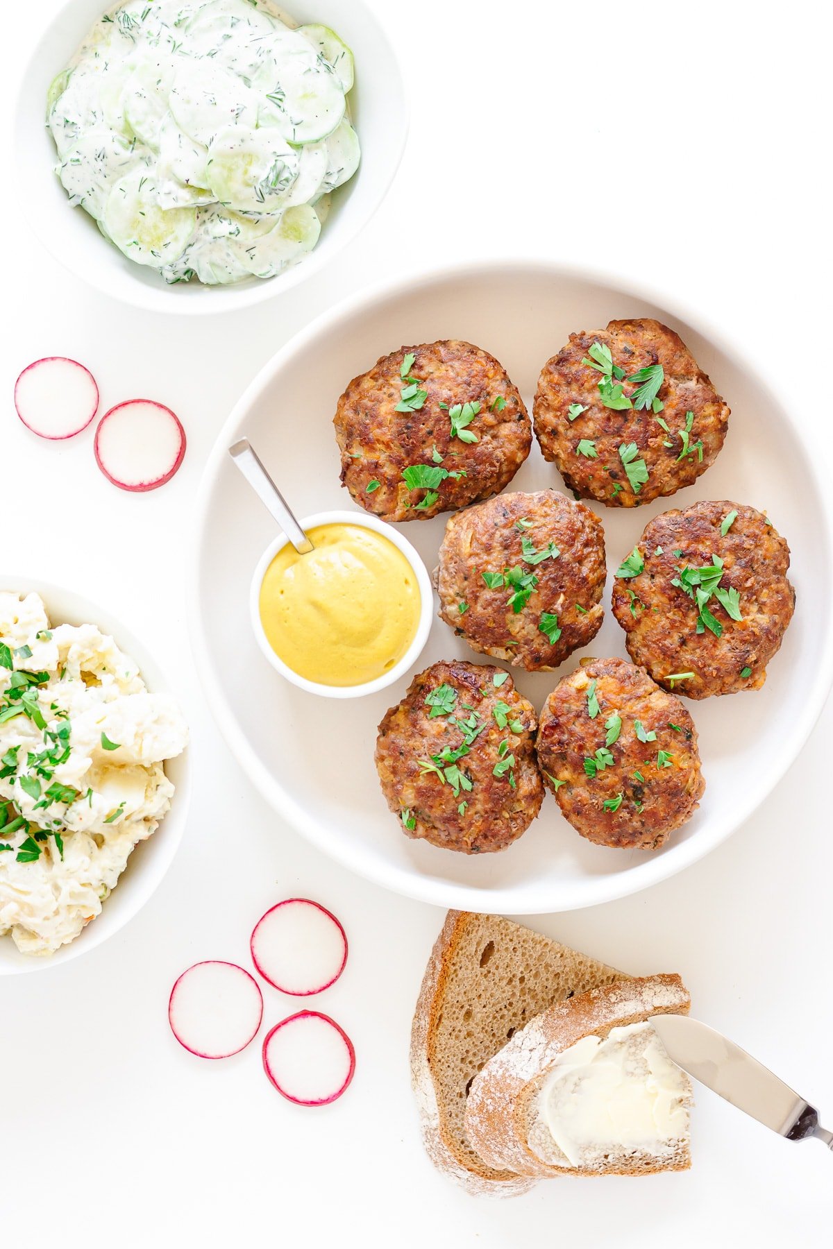 German frikadellen in a round white dish with mustard and scattering of chopped parsley.  Surrounded by cucumber salad, potato salad, German rye bread and radish slices.