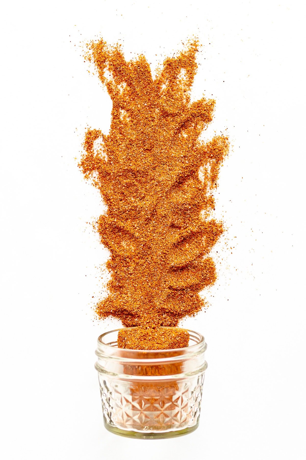 Small glass jar with fajita seasoning spilling out of it onto a white background.