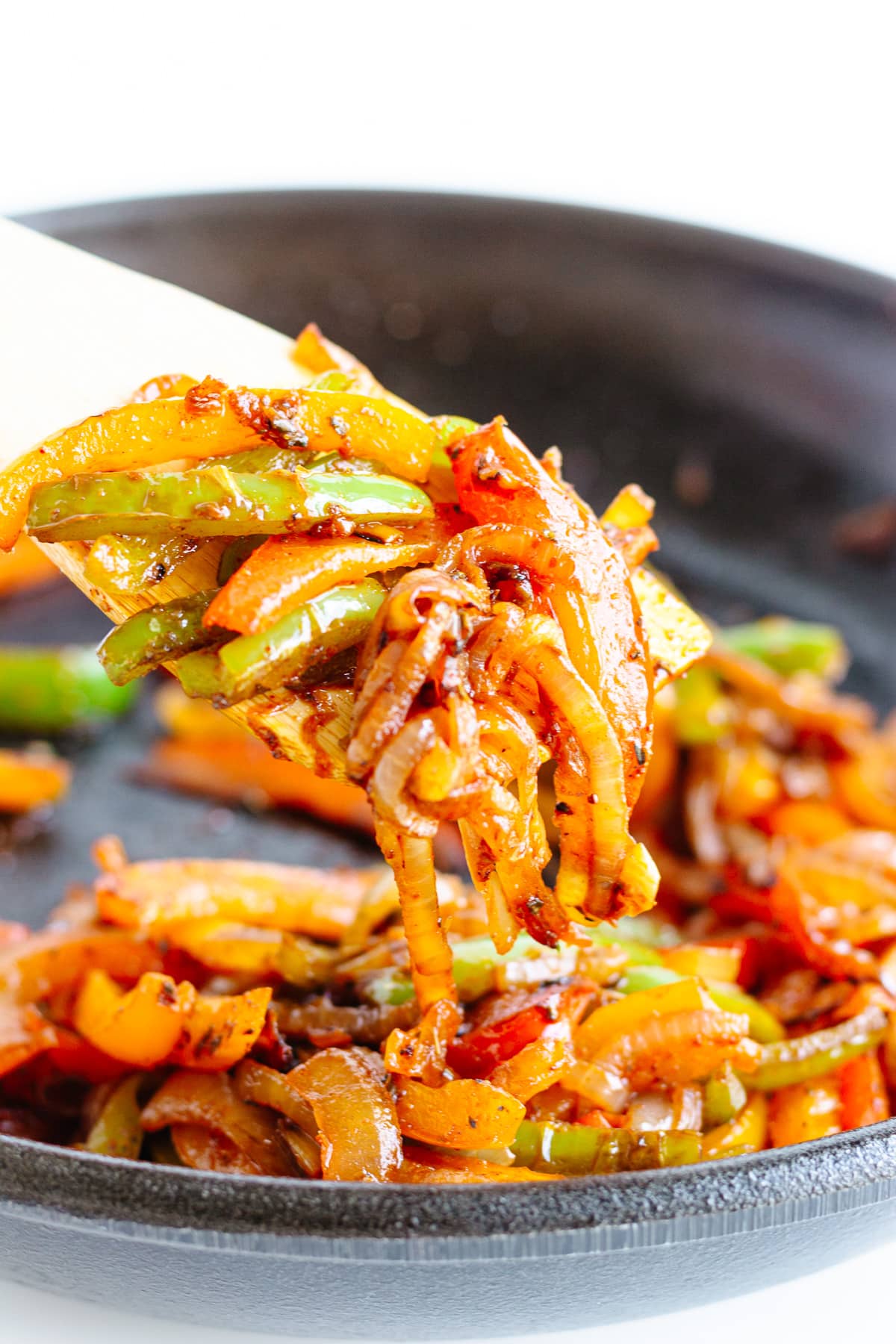 Wooden spoon lifting up some fajita vegetables from a cast iron skillet.