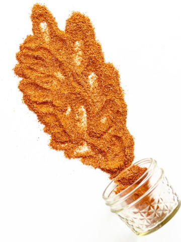 Small glass jar on its side with fajita seasoning spilling out across a white background.