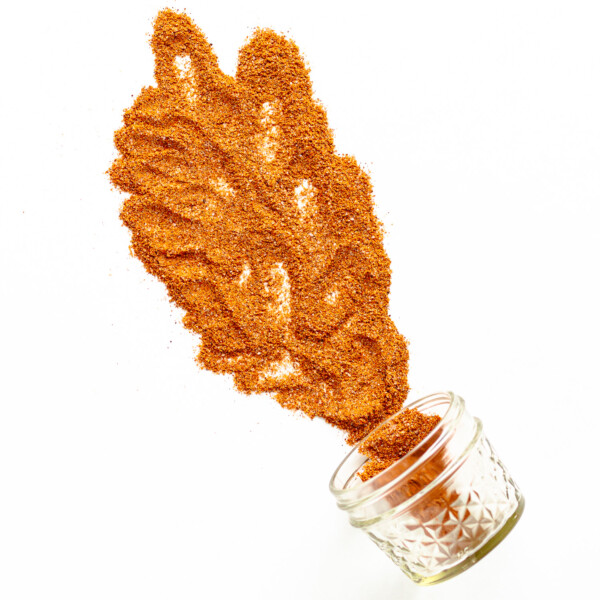 Small glass jar on its side with fajita seasoning spilling out across a white background.