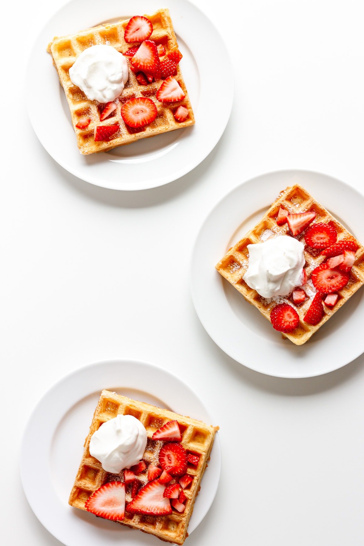 Three white plates each holding one square waffle topped with fresh strawberries and whipped cream.