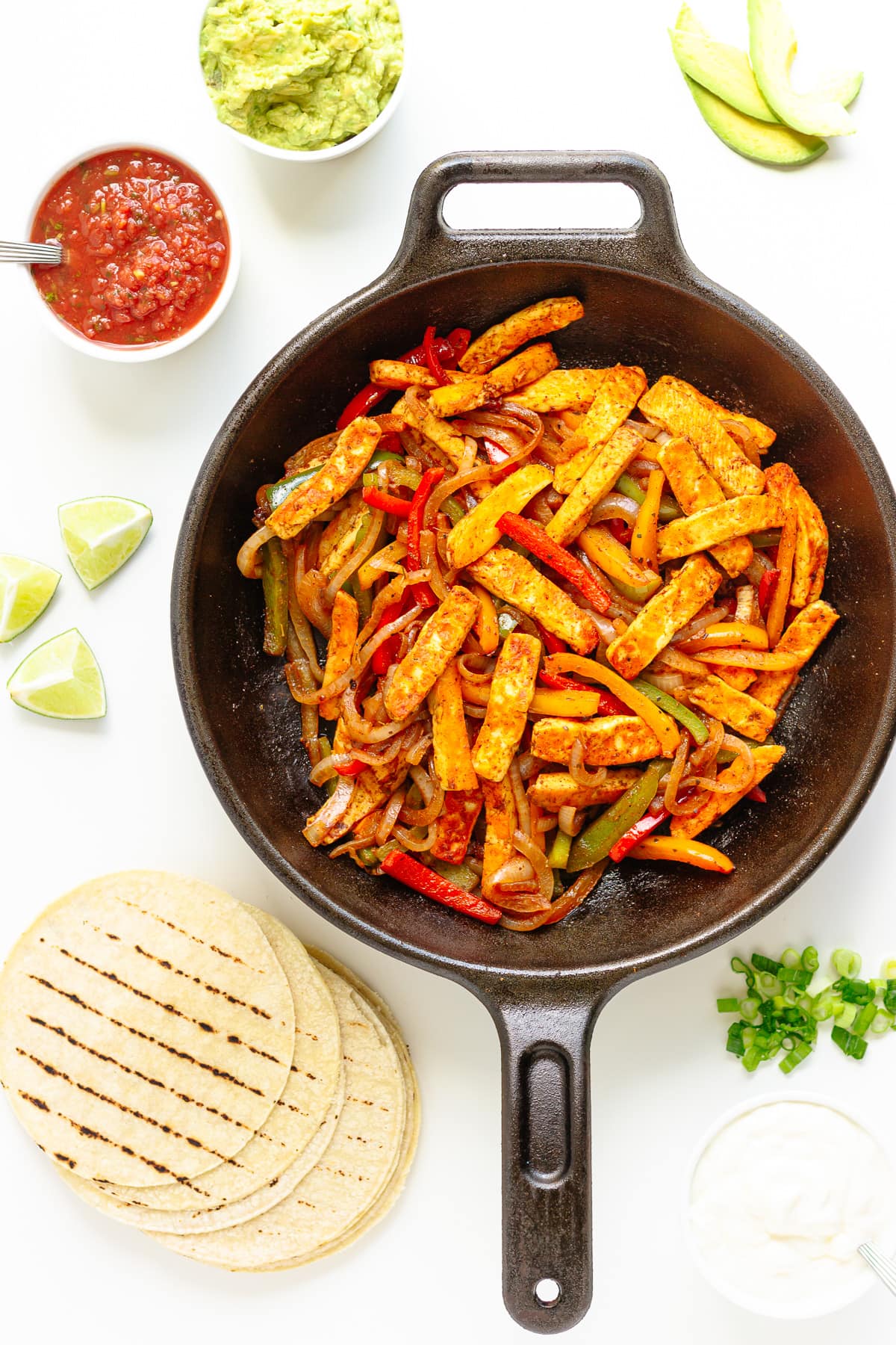 Halloumi veggie fajita mixture in a cast iron skillet with tortillas and a variety of toppings around it.