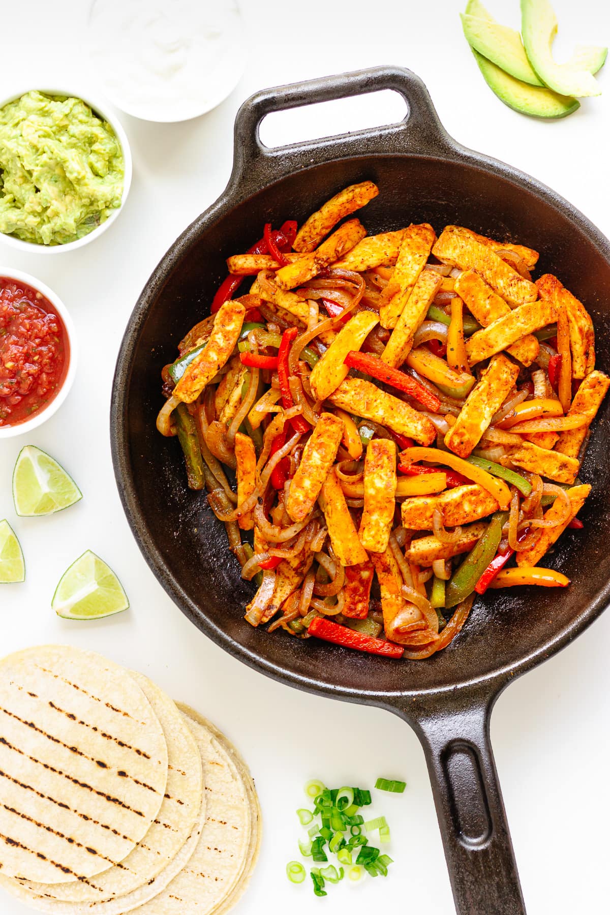 Flat lay of halloumi vegetable fajita mixture in a cast iron skillet surrounded by tortillas and toppings.