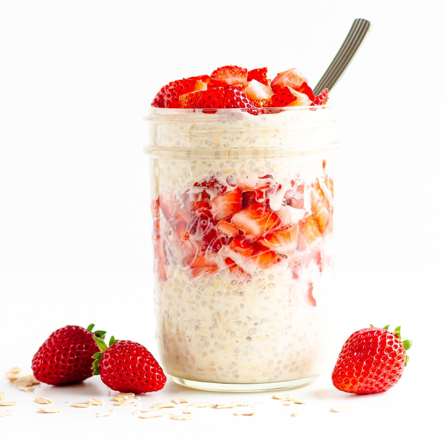 Jar containing layers of overnight oats and chopped strawberries with some whole strawberries and rolled oats scattered around it.