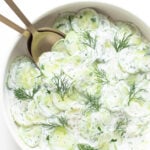 Close up of bowl containing creamy German cucumber salad (Gurkensalat) with gold salad servers.